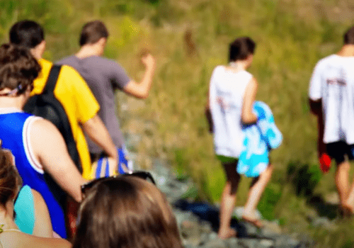 A group of people walking along a rocky path, some carrying towels and wearing casual summer clothing.