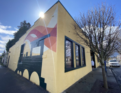 A colorful mural on a yellow building corner, featuring abstract shapes and a tree in front under a blue sky.