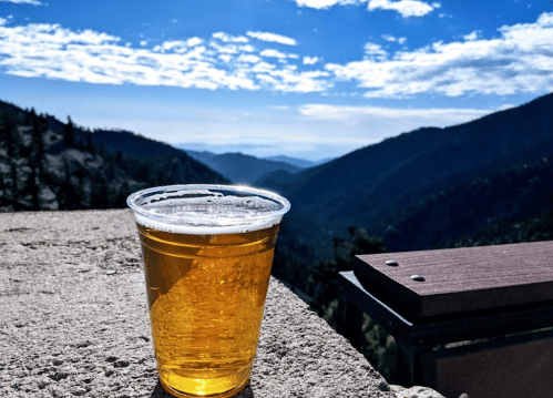 A clear plastic cup of beer sits on a stone ledge, overlooking a scenic mountain view under a blue sky.
