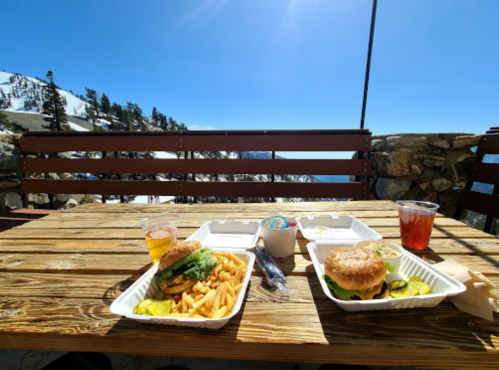 A sunny outdoor table with two burger meals, fries, and drinks, overlooking a snowy mountain landscape.