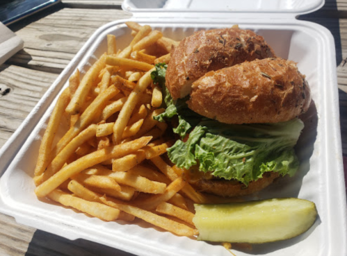 A sandwich with lettuce on a bun, served with a side of golden fries and a pickle, in a takeout container.