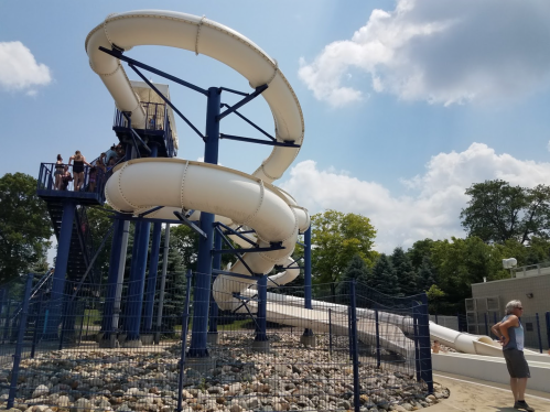 A water slide with a spiral design, surrounded by rocks and a fence, under a sunny sky with a person standing nearby.