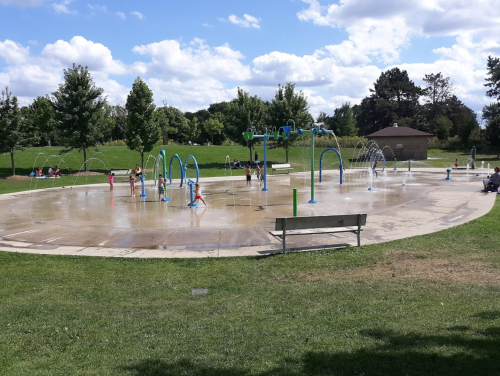 A splash pad in a park with children playing, surrounded by green grass and trees under a partly cloudy sky.