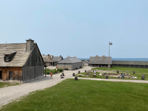 Historic buildings and a flag in a grassy area, with a pathway leading to a garden and the ocean in the background.