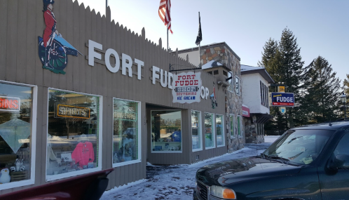 Exterior of a shop named "Fort Fudge," featuring signs for shirts, fudge, and ice cream, with snow on the ground.