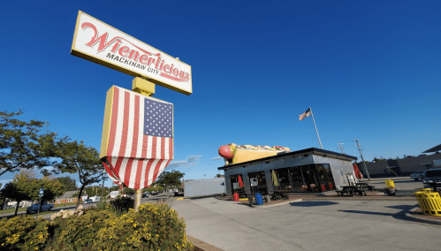 A colorful restaurant sign with an American flag, featuring a hot dog theme, in Mackinaw City under a clear blue sky.