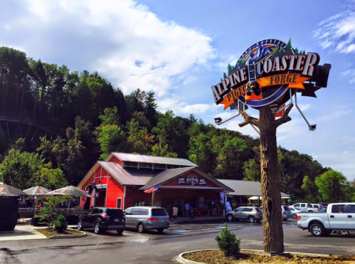A red building with a sign reading "Alpine Coaster" surrounded by trees and parked cars on a sunny day.