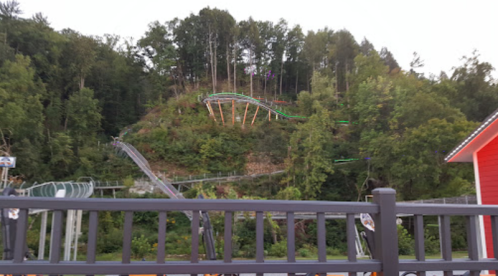 A roller coaster track winds through a wooded hillside, with trees surrounding the area and a red building in the foreground.