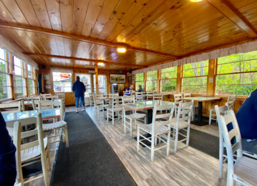 A cozy wooden interior of a restaurant with empty tables and a few patrons, surrounded by large windows.