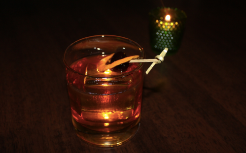 A glass of cocktail with a cherry and orange twist, illuminated by a soft candlelight in the background.