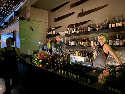 Two bartenders smile and pose behind a well-stocked bar, with bottles and tools visible in a dimly lit setting.