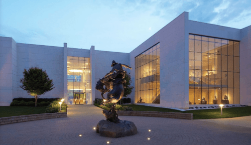 Modern building with large glass windows, featuring a bronze statue of a cowboy on horseback in a landscaped area.