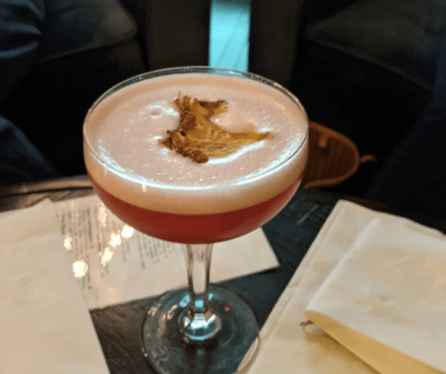 A cocktail in a stemmed glass, topped with a dried leaf, resting on a table with a napkin and menu in the background.