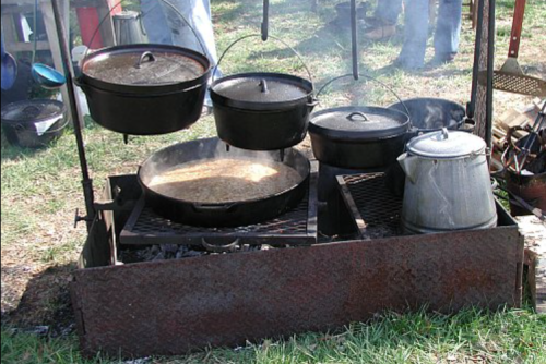 A rustic outdoor cooking setup with several black pots and a kettle hanging over a fire on a metal grate.