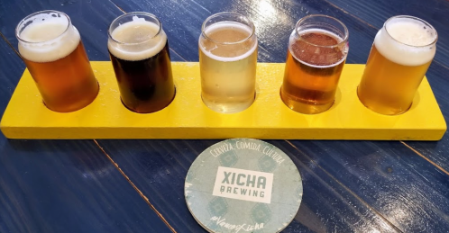 A flight of five craft beers on a yellow paddle, with a coaster from Xicha Brewing on a blue table.