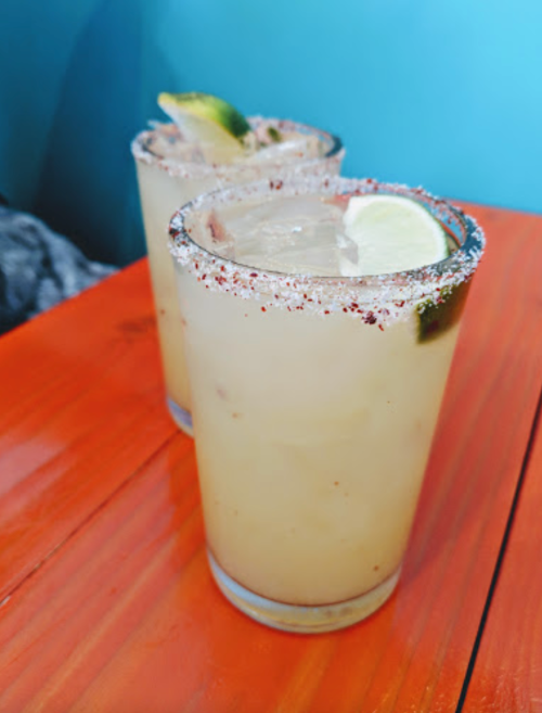 Two refreshing cocktails with lime slices and a salted rim, placed on a vibrant orange table.