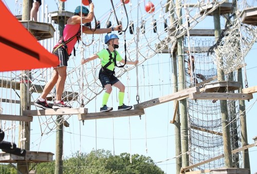 A child in a bright outfit navigates a ropes course while an adult assists from above.