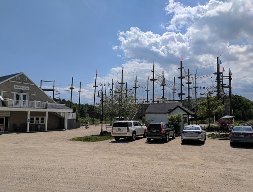A parking area with several cars, a building, and a large outdoor ropes course in the background under a cloudy sky.