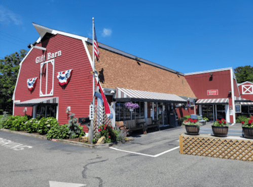 A red barn-style gift shop with flower planters and American flags, set against a clear blue sky.