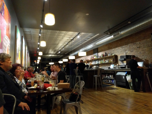 A lively restaurant interior with patrons dining, a bar area, and colorful wall art. Warm lighting creates a cozy atmosphere.