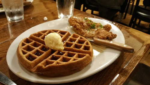 A plate with a golden waffle topped with butter and syrup, alongside a piece of fried chicken.
