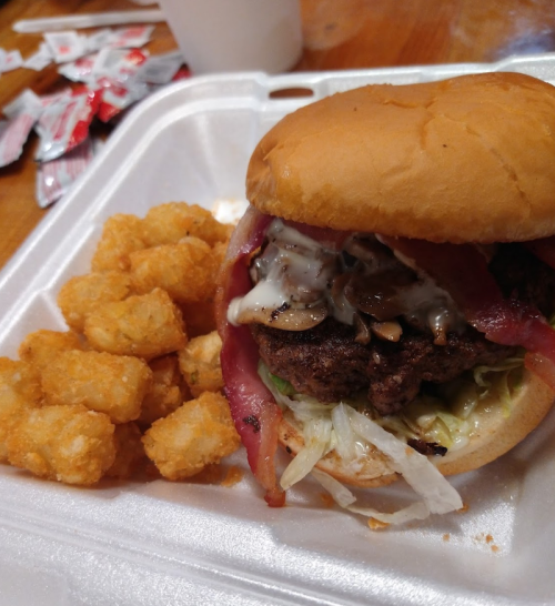 A juicy burger with lettuce, tomato, and sauce, served with a side of crispy tater tots in a takeout container.