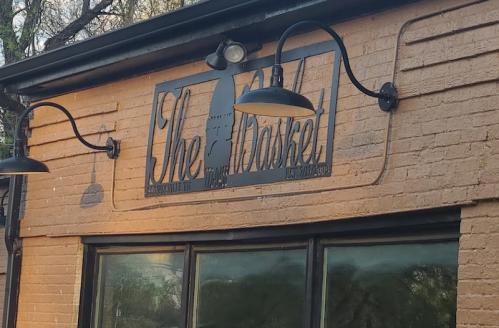 Sign for "The Basket" restaurant, featuring a decorative design and outdoor lighting on a brick wall.