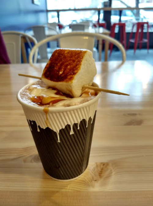 A cup of hot chocolate topped with a toasted marshmallow on a stick, placed on a wooden table.