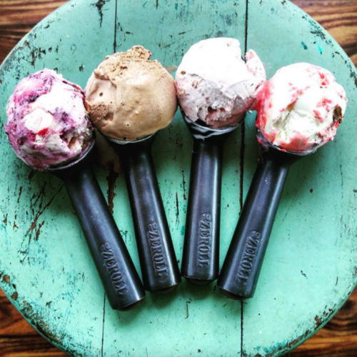 Four scoops of ice cream in black scoops on a rustic green wooden surface. Flavors include chocolate, berry, and strawberry.