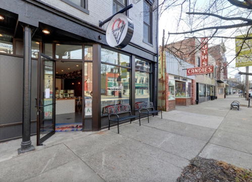 Exterior view of a candy shop with a sign reading "Aunt's Candies," featuring benches and a sidewalk in a quaint neighborhood.
