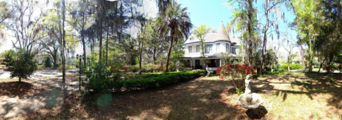 A panoramic view of a historic house surrounded by lush greenery and colorful flowers under a clear blue sky.