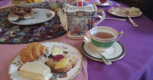 A plate with a croissant, sandwich, and cookie beside a teacup and teapot on a colorful table setting.