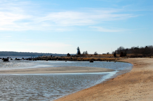 A serene beach scene with gentle waves, a sandy shore, and a distant lighthouse under a clear blue sky.