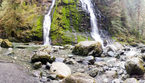 A serene waterfall cascades over moss-covered rocks, surrounded by lush greenery and a rocky riverbed.