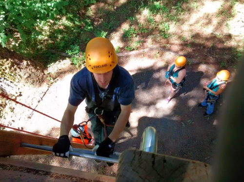 A person in a helmet climbs a wooden structure while two others watch from below, also wearing helmets.