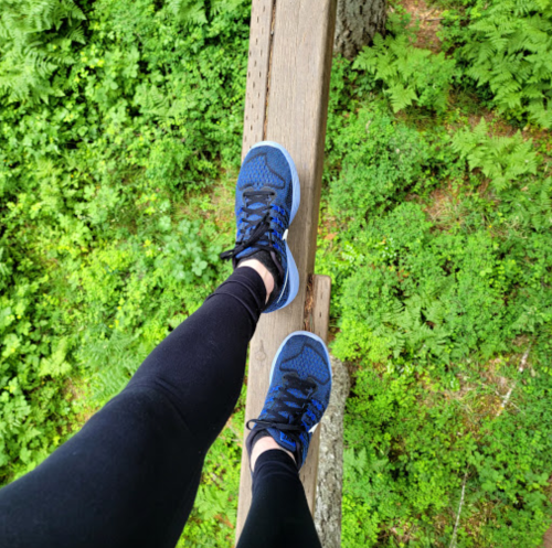 A person stands on a wooden beam, wearing black leggings and blue sneakers, surrounded by lush green foliage.