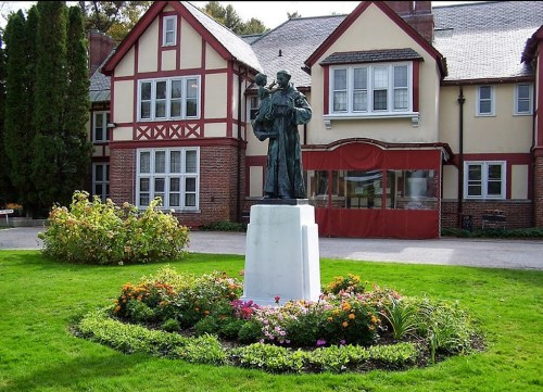 A statue stands on a pedestal in a garden, surrounded by colorful flowers, in front of a large, traditional-style house.