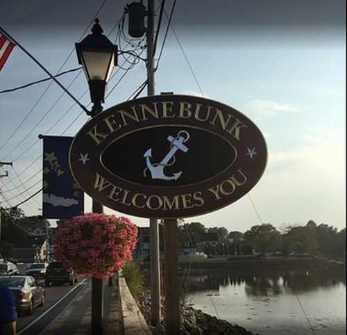 Sign reading "Kennebunk Welcomes You" with an anchor design, surrounded by flowers and a scenic waterfront view.