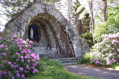 A stone archway shrine surrounded by blooming pink rhododendrons and greenery, featuring a statue of a figure inside.