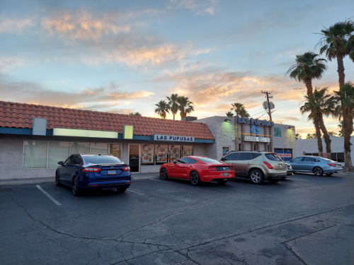 A parking lot with three cars in front of a restaurant building featuring a sign for "Las Pupusas" and palm trees.