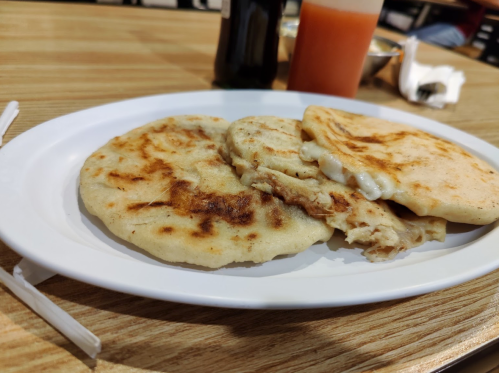 Three golden-brown pupusas on a white plate, with a bottle of sauce and a drink in the background.