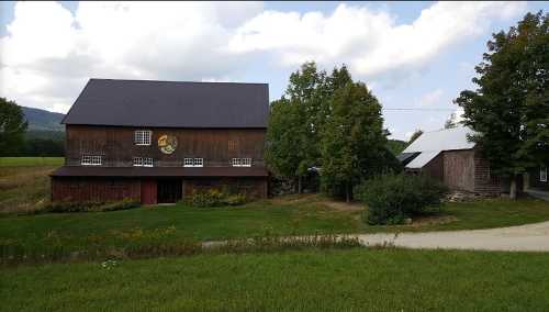 A rustic wooden barn with a circular emblem, surrounded by green grass and trees under a cloudy sky.