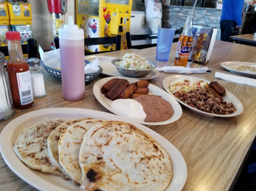 A table with plates of food, including tortillas, beans, coleslaw, fried plantains, and drinks in a casual dining setting.