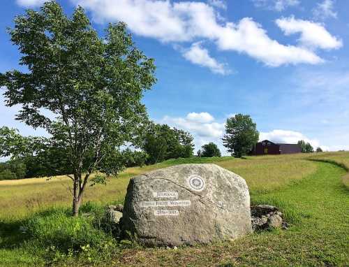 A large rock with a plaque stands in a grassy field, with a house and trees in the background under a blue sky.