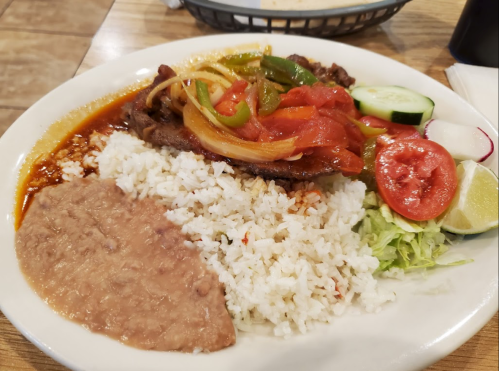 A plate of rice, refried beans, grilled meat with peppers and tomatoes, and fresh vegetables including lettuce and lime.