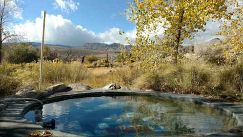 A serene hot spring surrounded by autumn foliage and mountains under a partly cloudy sky.