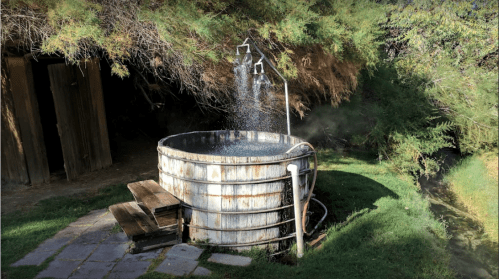 A rustic wooden hot spring tub with water flowing from two spouts, surrounded by greenery and a stone pathway.