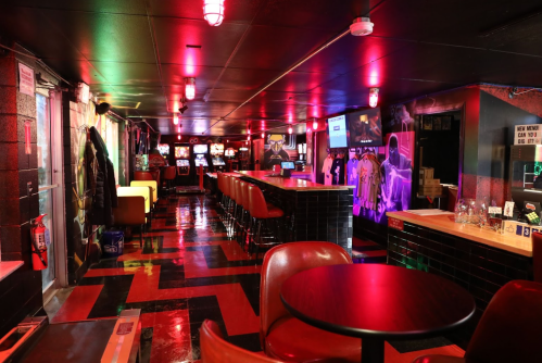 A dimly lit arcade bar with colorful lights, a long bar, and gaming machines lining the walls.