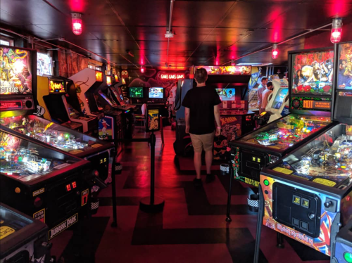A dimly lit arcade filled with pinball machines and retro games, with a person walking through the aisles.