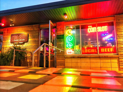 A brightly lit arcade entrance with neon signs, featuring "Coin Slot" and "Local Beef," set against a colorful evening sky.